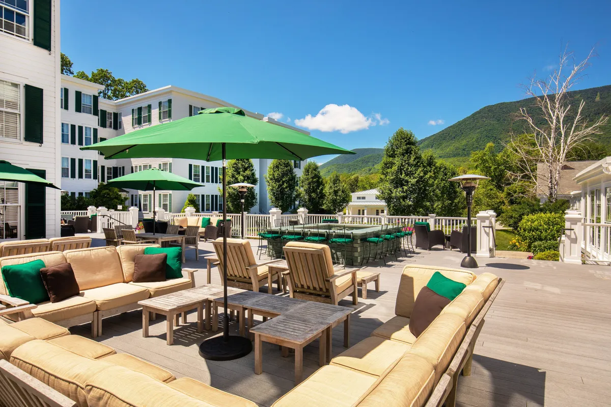 An inviting wooden deck lined with green umbrellas and comfortable lounge seating faces a sweeping mountain view. Guests can relax, socialize, or dine while soaking up the scenery, illustrating the resort’s blend of activity and relaxation. The scene captures the transition from active pursuits to restful moments.