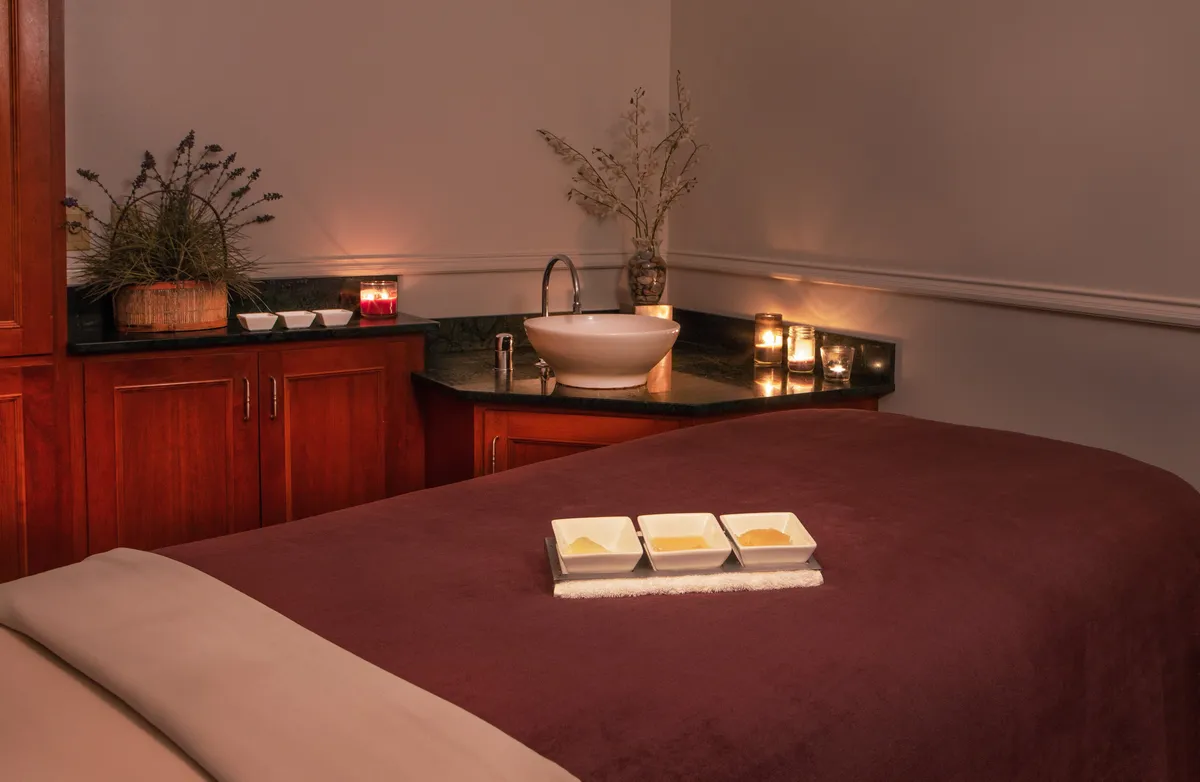 A serene spa treatment room featuring a comfortable massage table, a bowl with essential oils, candles, a sink, and a decorative plant. The setting is intimate and tranquil, with soft lighting creating an atmosphere ideal for rejuvenation and wellness treatments.