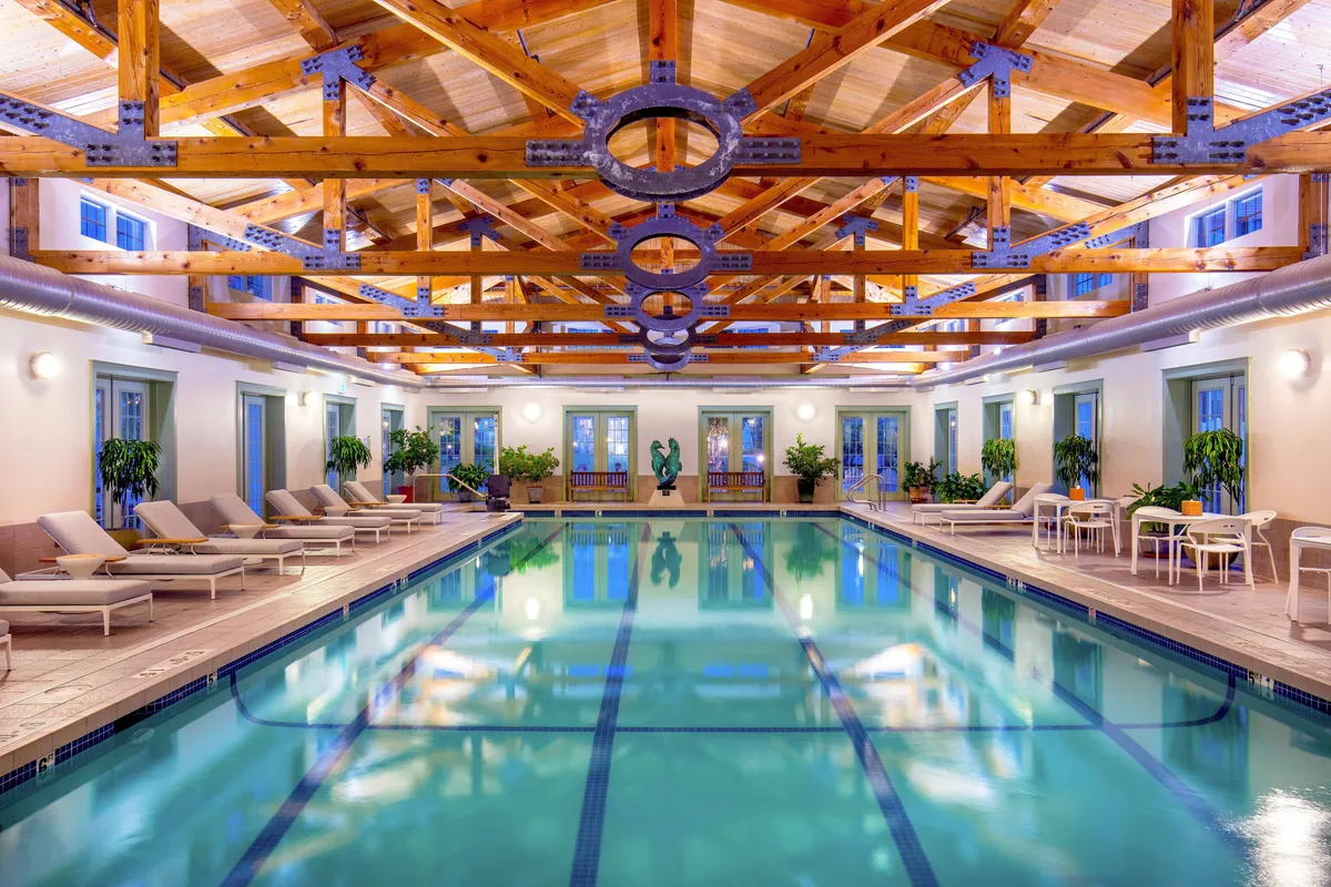 This image features a luxurious indoor swimming pool area with exposed wooden beams, elegant lounge chairs, and decorative plants. The pool is inviting and serene, with natural light filtering in, suggesting a calming space ideal for relaxation or gentle exercise. The presence of nearby dining and lounge areas hints at an all-encompassing wellness environment.
