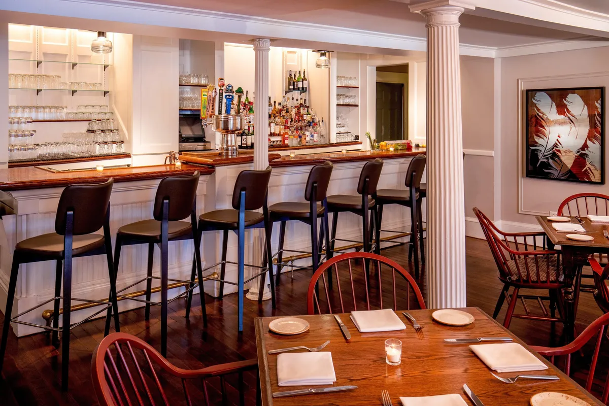 A warmly lit interior dining space featuring a polished bar counter, comfortable stools, elegant dining tables, and thoughtfully curated artwork adorning the walls. The scene exudes a friendly, relaxed ambiance with guests engaged in lively conversation and enjoying drinks and small plates.