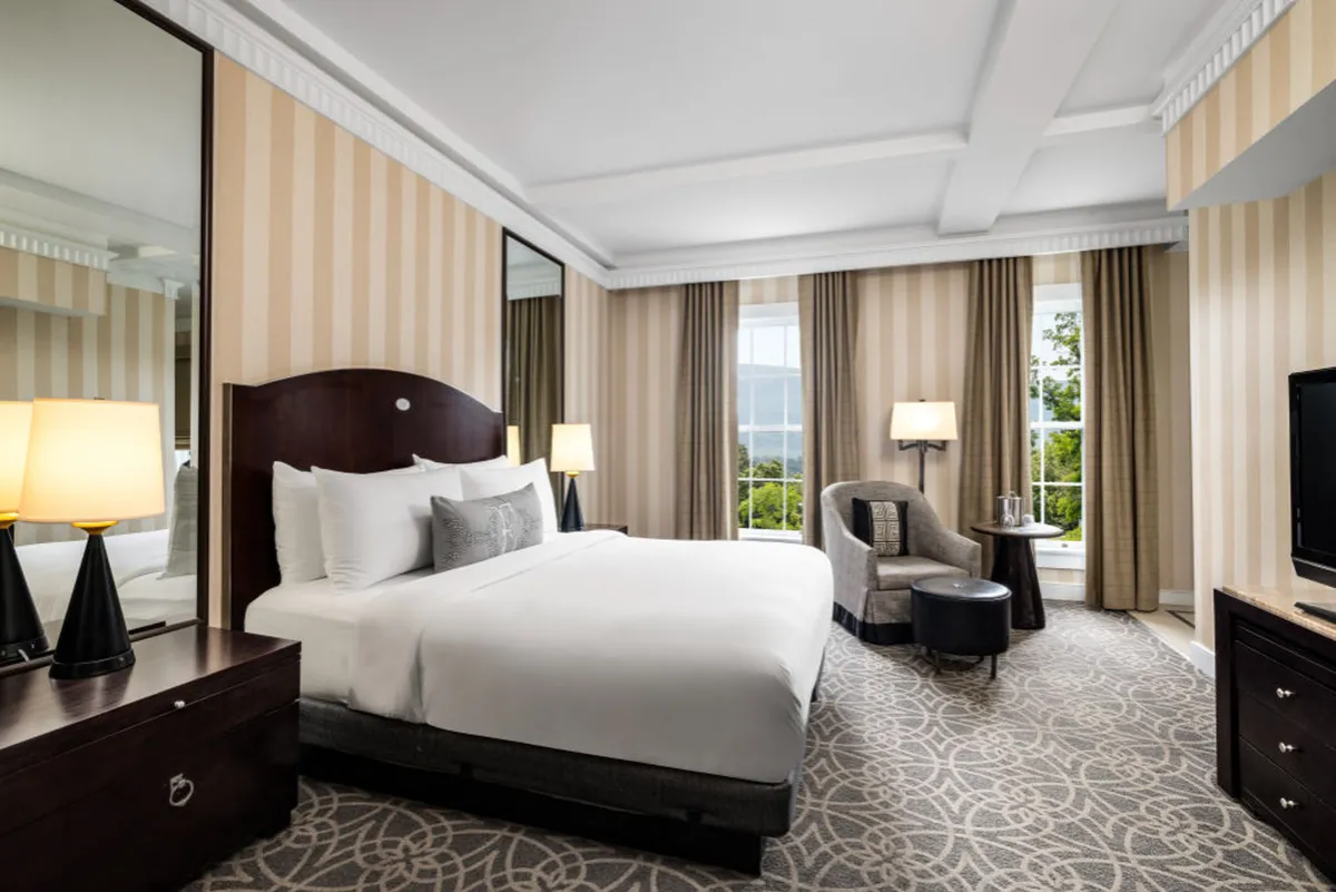 The image showcases a bright, elegantly furnished studio interior with a plush bed, cozy armchair positioned by a large window letting in abundant natural light, a classic nightstand, and a wall-mounted television. The scene radiates warmth, comfort, and an inviting atmosphere perfect for unwinding after a day at the resort.