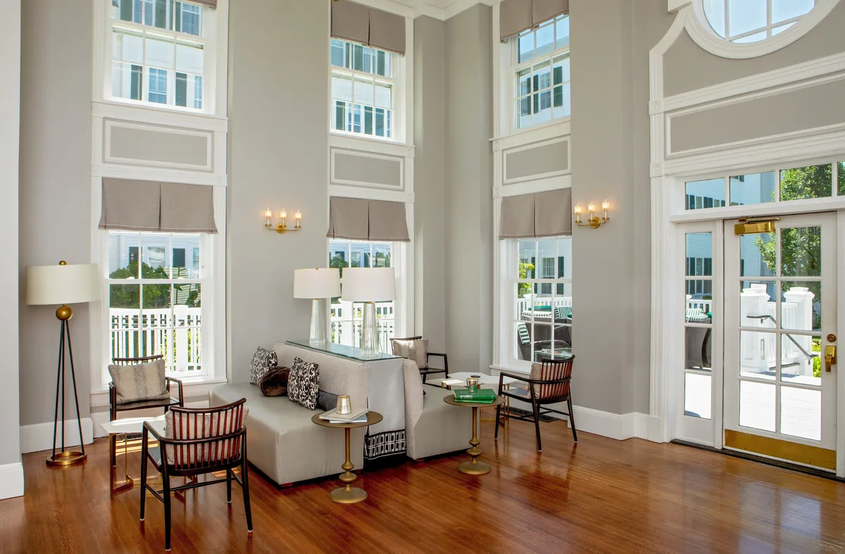A sunlit interior lounge area featuring large windows that flood the space with natural light. The room contains a comfortable sofa, a coffee table, and a floor lamp, arranged to create a welcoming and cozy seating nook. The soothing neutral palette and refined decor evoke classic New England style while supporting quiet relaxation or thoughtful work.