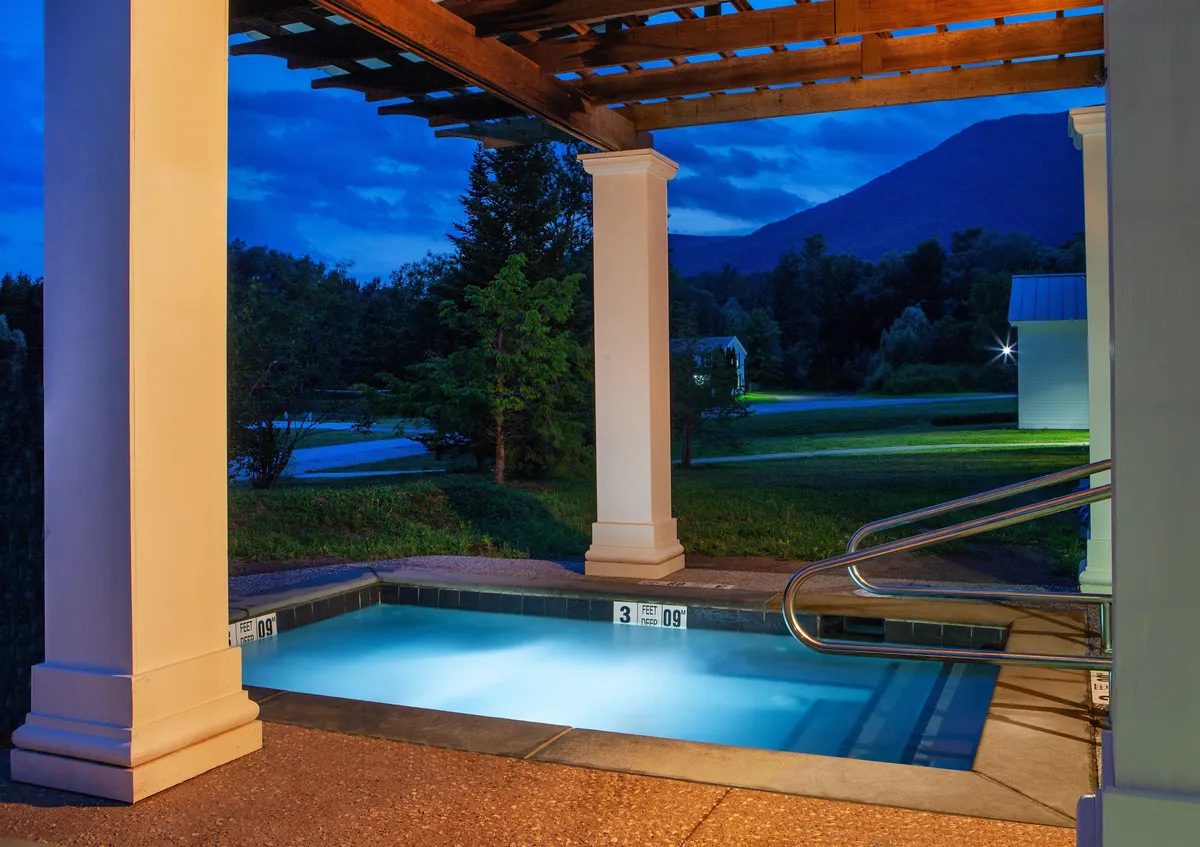 This image showcases the exterior of the resort at night, highlighting an illuminated pool beneath a wooden pergola, surrounded by a tranquil landscape. The soft lighting and serene atmosphere create a romantic backdrop, inviting guests to relax, swim, or stargaze together. The scene subtly hints at the possibility of sharing stories poolside, capturing the peaceful and intimate mood ideal for couples.