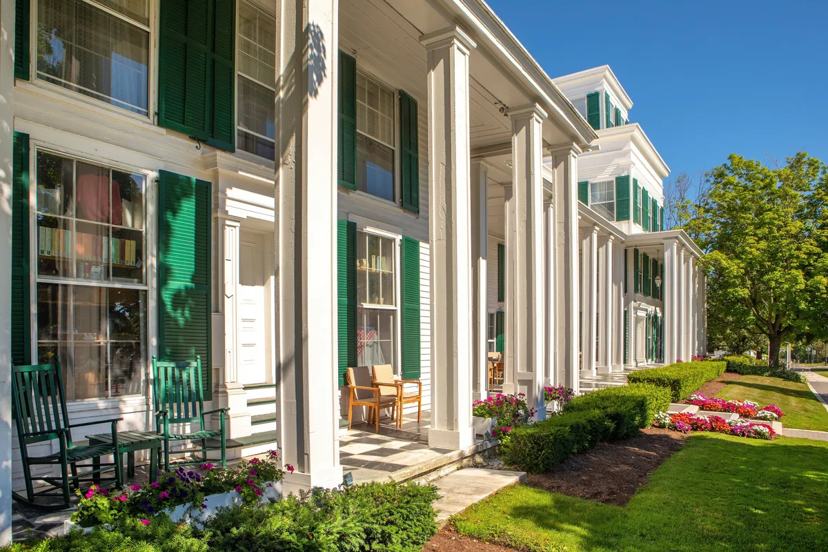 A classic exterior porch with large columns, green shutters, and inviting rocking chairs overlooking vibrant flower beds. The setting suggests a peaceful spot for enjoying morning coffee or socializing with neighbors against a backdrop of New England charm.